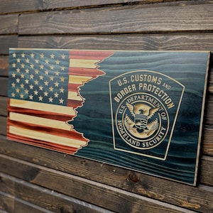 Könnte beinhalten: Ein Holzschild mit einem abgenutzten amerikanischen Flaggen-Design und dem Siegel der US-Zoll- und Grenzschutzbehörde. Das Siegel ist in Gold gehalten und zeigt einen Adler mit einem Schild und den Worten "U.S. Department of Homeland Security".