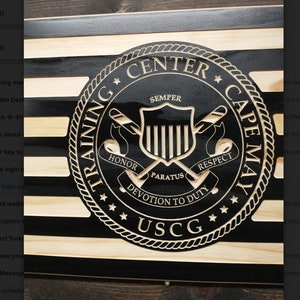 Op de afbeelding: Een zwart-wit houten bord met een zwart-wit embleem van de Amerikaanse kustwacht. Het embleem toont een schild met de woorden "Eer", "Semper", "Paratus", "Respect" en "Toewijding aan de plicht". De woorden "Training Center Cape May" staan rond het embleem.