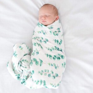 K&ouml;nnte beinhalten: Ein schlafendes Neugeborenes, eingewickelt in eine wei&szlig;e Musselin-Decke. Die Decke hat ein Aquarell-Eukalyptusblatt-Design in Gr&uuml;nt&ouml;nen. Das Baby liegt auf einem wei&szlig;en Laken.