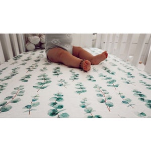 K&ouml;nnte beinhalten: Wei&szlig;es Kinderbettlaken mit einem sich wiederholenden Muster aus gr&uuml;nen Eukalyptusbl&auml;ttern. Ein Baby in einem grauen Strampler sitzt auf dem Laken. Das Kinderbett hat wei&szlig;e Gitterst&auml;be.