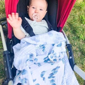 K&ouml;nnte beinhalten: Ein Baby in einem roten Kinderwagen winkt. Das Baby tr&auml;gt ein gestreiftes Oberteil und hat einen Schnuller. Eine blau-wei&szlig;e Blumendecke bedeckt das Baby. Der Kinderwagen hat einen schwarzen Rahmen und steht auf Gras.