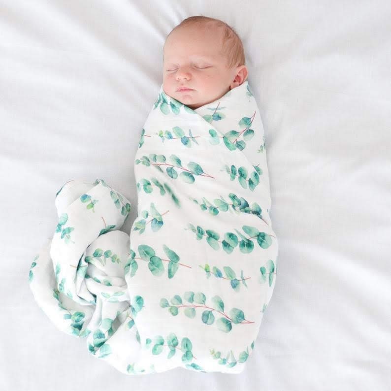 K&ouml;nnte beinhalten: Ein schlafendes Neugeborenes, eingewickelt in eine wei&szlig;e Musselin-Decke. Die Decke hat ein Aquarell-Eukalyptusblatt-Design in Gr&uuml;nt&ouml;nen. Das Baby liegt auf einem wei&szlig;en Laken.