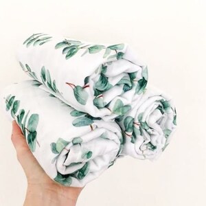 K&ouml;nnte beinhalten: Drei gerollte wei&szlig;e Decken mit einem gr&uuml;nen Eukalyptusblattmuster. Die Decken sind weich und scheinen aus einem leichten Stoff zu bestehen. Die Bl&auml;tter haben einen Aquarelleffekt mit Gr&uuml;nt&ouml;nen und roten Stielen.