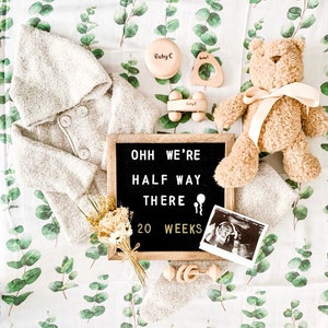 K&ouml;nnte beinhalten: Eine Baby-Ank&uuml;ndigung mit einem grauen Kapuzenj&auml;ckchen, einem Teddyb&auml;ren, Holzspielzeug und einer Buchstaben-Tafel mit Text. Die Gegenst&auml;nde liegen auf einer Decke mit Eukalyptusmuster. Auch ein Ultraschallbild ist zu sehen.