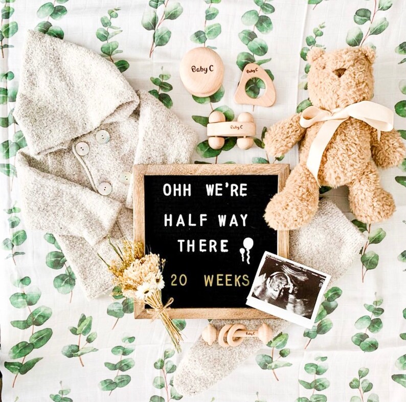 K&ouml;nnte beinhalten: Eine Baby-Ank&uuml;ndigung mit einem grauen Kapuzenj&auml;ckchen, einem Teddyb&auml;ren, Holzspielzeug und einer Buchstaben-Tafel mit Text. Die Gegenst&auml;nde liegen auf einer Decke mit Eukalyptusmuster. Auch ein Ultraschallbild ist zu sehen.