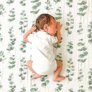 K&ouml;nnte beinhalten: Ein Baby in einem wei&szlig;en Body liegt auf einer wei&szlig;en Decke mit einem gr&uuml;nen Eukalyptusblattmuster. Das Baby liegt auf der Seite, Arme und Beine sind leicht angewinkelt. Die Decke hat ein weiches, strukturiertes Aussehen.