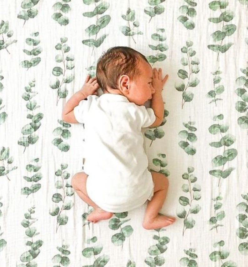 K&ouml;nnte beinhalten: Ein Baby in einem wei&szlig;en Body liegt auf einer wei&szlig;en Decke mit einem gr&uuml;nen Eukalyptusblattmuster. Das Baby liegt auf der Seite, Arme und Beine sind leicht angewinkelt. Die Decke hat ein weiches, strukturiertes Aussehen.