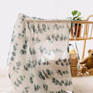 K&ouml;nnte beinhalten: Eine wei&szlig;e Babydecke mit einem sich wiederholenden Muster aus Eukalyptusbl&auml;ttern in Gr&uuml;n- und Blaut&ouml;nen. Die Decke ist &uuml;ber einen Holzrahmen drapiert. Im Hintergrund sind eine kleine Topfpflanze, ein Teddyb&auml;r und Weidenk&ouml;rbe zu sehen. Das Material ist leicht.