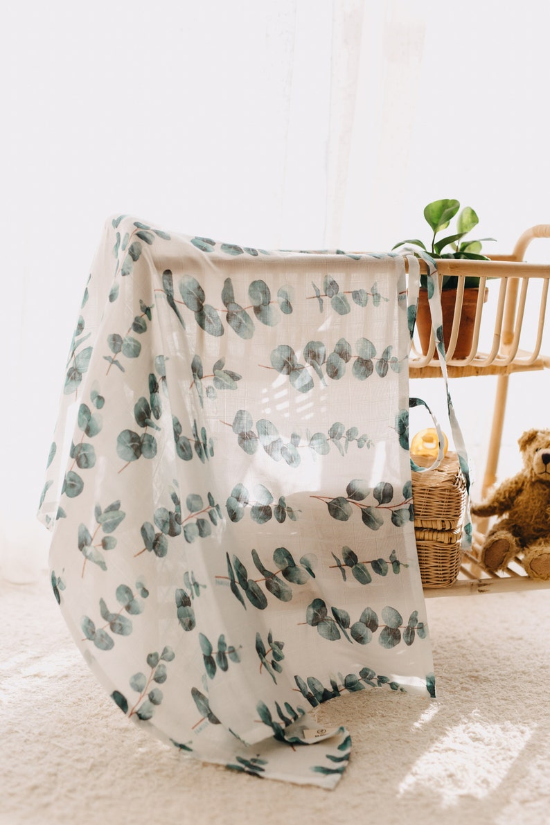 K&ouml;nnte beinhalten: Eine wei&szlig;e Babydecke mit einem sich wiederholenden Muster aus Eukalyptusbl&auml;ttern in Gr&uuml;n- und Blaut&ouml;nen. Die Decke ist &uuml;ber einen Holzrahmen drapiert. Im Hintergrund sind eine kleine Topfpflanze, ein Teddyb&auml;r und Weidenk&ouml;rbe zu sehen. Das Material ist leicht.