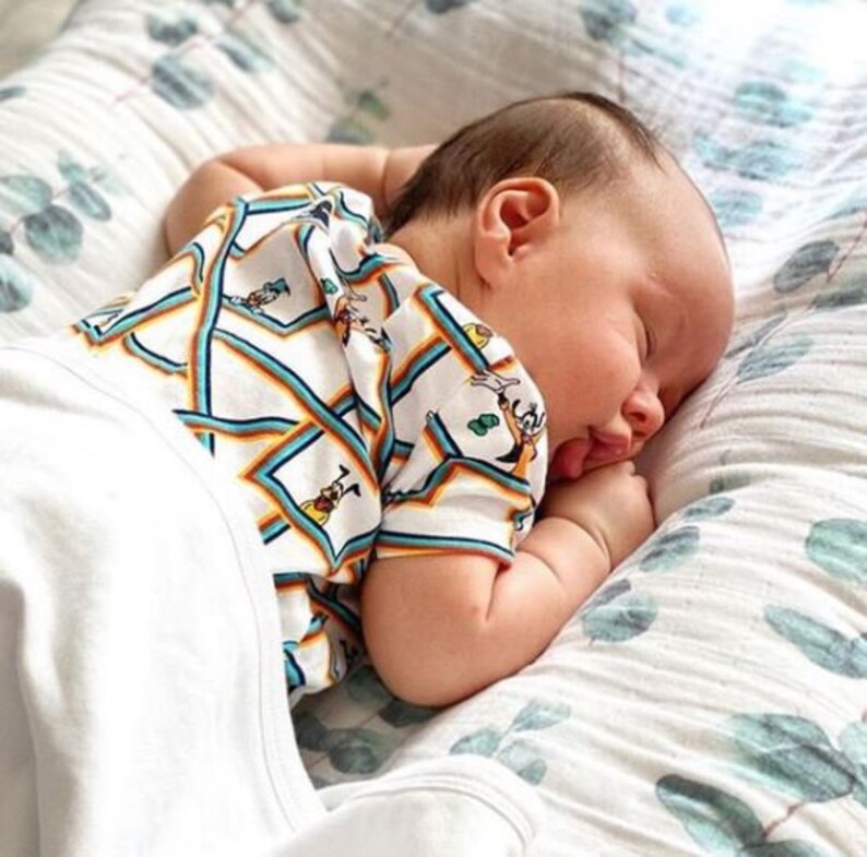 K&ouml;nnte beinhalten: Ein schlafendes Baby in einem wei&szlig;en Strampler mit einem bunten Muster aus Zeichentrickfiguren und geometrischen Formen. Das Baby liegt auf einer gemusterten Decke mit einer weichen, neutralen Farbpalette.