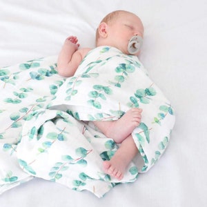 K&ouml;nnte beinhalten: Ein schlafendes Baby, eingewickelt in eine wei&szlig;e Musselin-Decke mit einem gr&uuml;nen Eukalyptusblattmuster. Das Baby tr&auml;gt einen Schnuller und liegt auf einer wei&szlig;en Oberfl&auml;che.