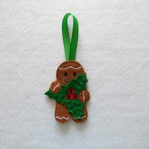 May include: A gingerbread man Christmas ornament with a green holly sprig and red berries. The ornament has a green ribbon for hanging.