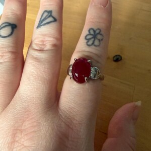 May include: A silver ring featuring a large, oval, deep red gemstone. The ring is worn on a finger with tattoos of a paper aeroplane, a flower, and other designs. The background is a light-coloured wooden surface.