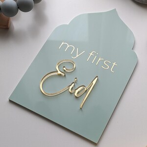 May include: A light blue sign with rounded edges and a pointed top, shaped like an arch. The sign says "my first Eid" in gold lettering.
