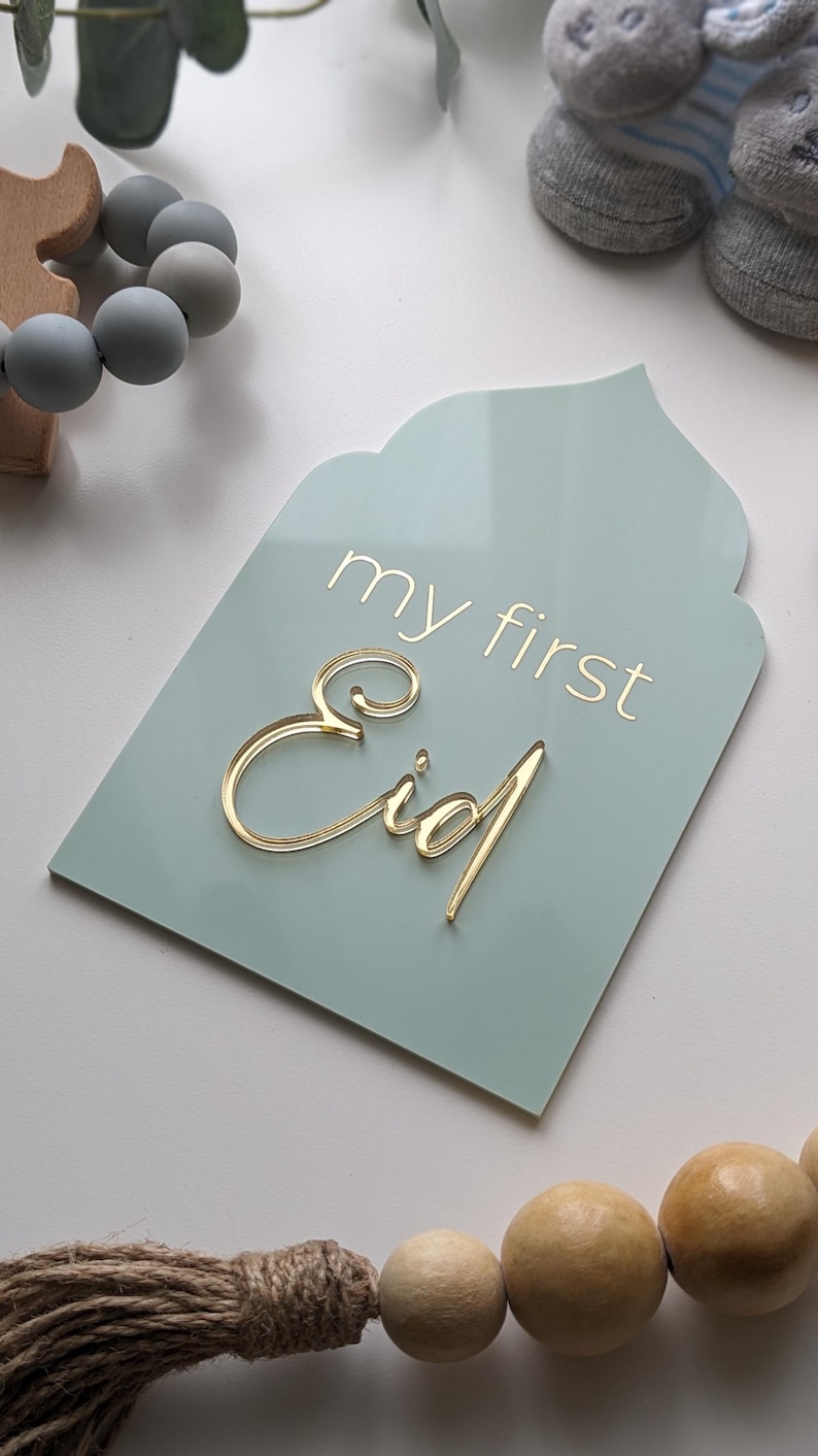 May include: A light blue sign with rounded edges and a pointed top, shaped like an arch. The sign says "my first Eid" in gold lettering.