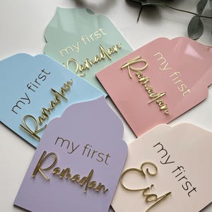May include: Four pastel-coloured wooden signs with the text "my first Ramadan" and "my first Eid" in gold lettering. The signs are shaped like arches and have a decorative design.