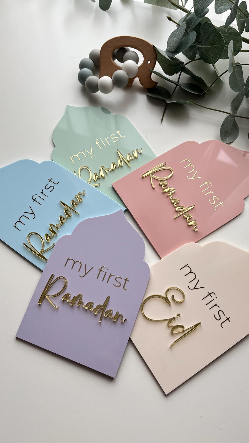 May include: Four pastel-coloured wooden signs with the text "my first Ramadan" and "my first Eid" in gold lettering. The signs are shaped like arches and have a decorative design.