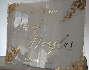 May include: A clear acrylic wedding sign with gold leaf accents. The sign reads "Welcome to the wedding of Sarah & Douglas October 19, 2023".