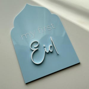 May include: A light blue sign with a white arch at the top. The sign says "my first Eid" in white lettering.