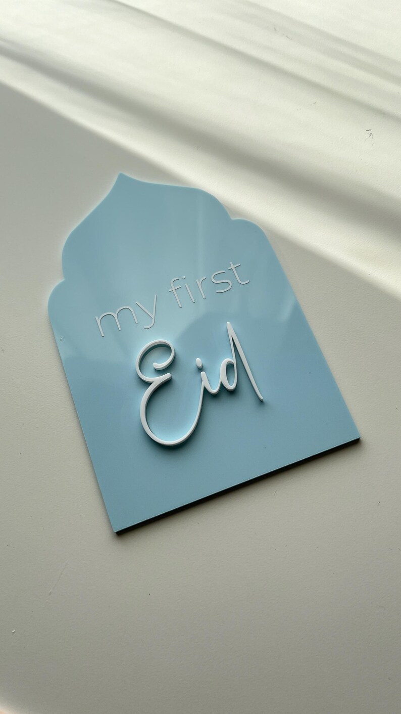 May include: A light blue sign with a white arch at the top. The sign says "my first Eid" in white lettering.