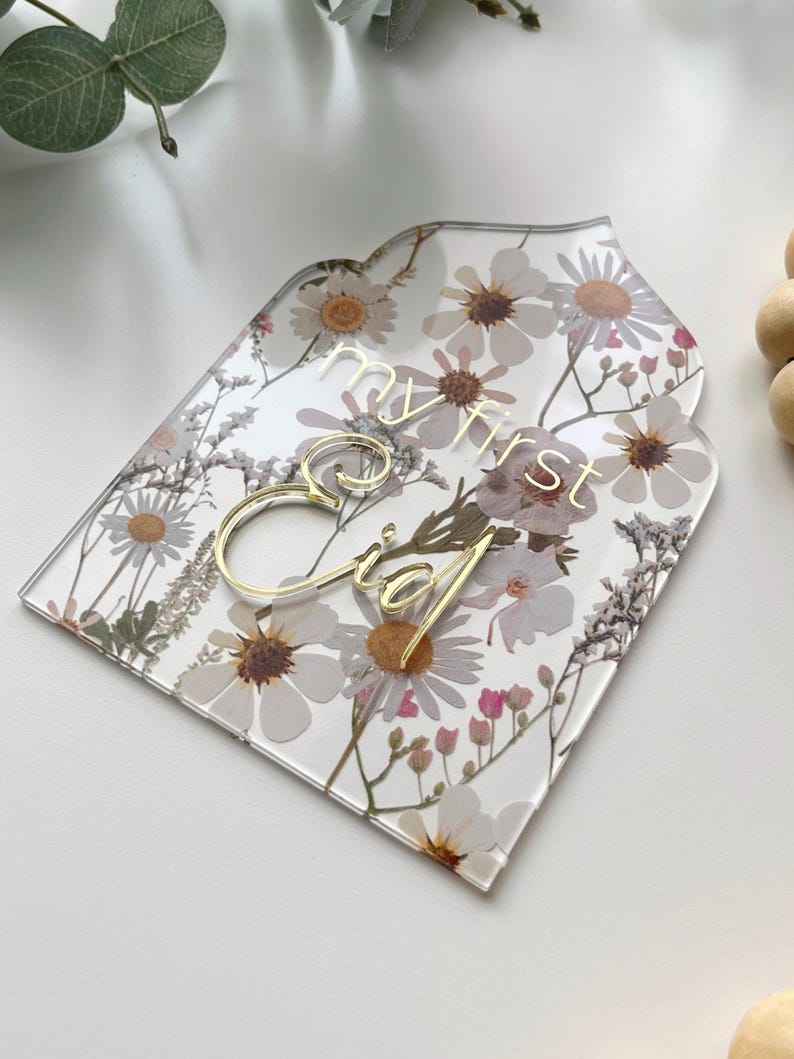 May include: A clear acrylic sign with a floral pattern and gold lettering that says "my first Eid".