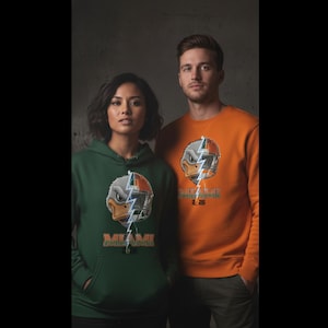 Pode incluir: Um moletom com capuz verde escuro e um moletom laranja. Ambas as peças de roupa apresentam um gráfico de um personagem semelhante a um pato com um raio e a palavra "MIAMI". O moletom laranja também tem o texto "2-26".