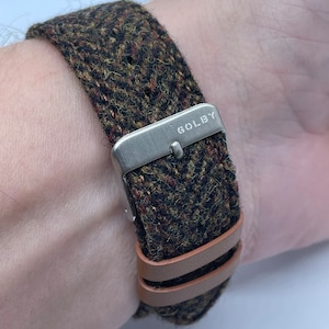 Puede incluir: Una correa de reloj de tweed marrón y verde con forro de cuero marrón y una hebilla plateada que dice "GOLBY".