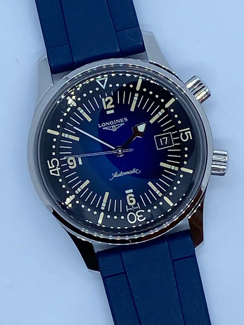 May include: A silver Longines watch with a blue face and a blue rubber strap. The watch has a date window at 3 o'clock and the words "Longines" and "Automatic" printed on the face.
