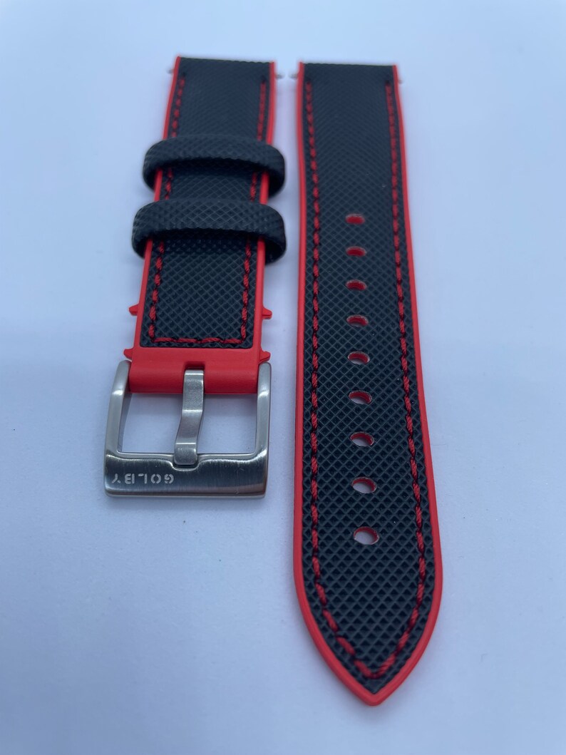 Puede incluir: Correa de reloj negra con costuras rojas y una hebilla plateada. La hebilla tiene la palabra "GOLIA" grabada.