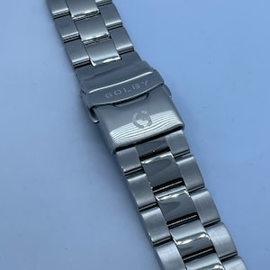 May include: A silver-coloured watch strap with a folding clasp. The strap is made up of numerous rectangular links. The clasp is engraved with the word "GOLBY" and a stylized "G" logo. The watch strap is designed for durability and style.