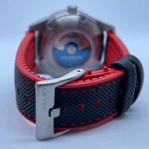 Puede incluir: Una correa de reloj negra y roja con una hebilla plateada. La hebilla tiene la palabra "GOLBY" grabada.