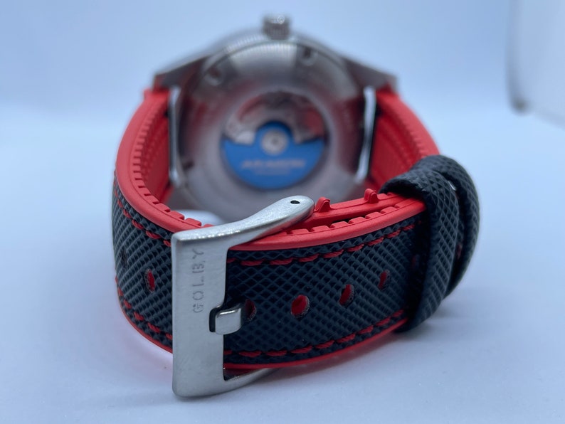 Puede incluir: Una correa de reloj negra y roja con una hebilla plateada. La hebilla tiene la palabra "GOLBY" grabada.