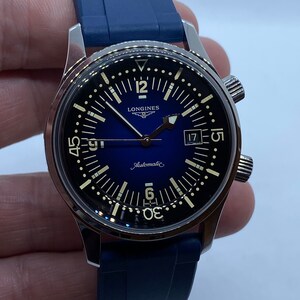 May include: A silver Longines watch with a blue face and a black rubber strap. The watch has a date window at the 3 o'clock position and the words "Automatic" and "Longines" printed on the face.