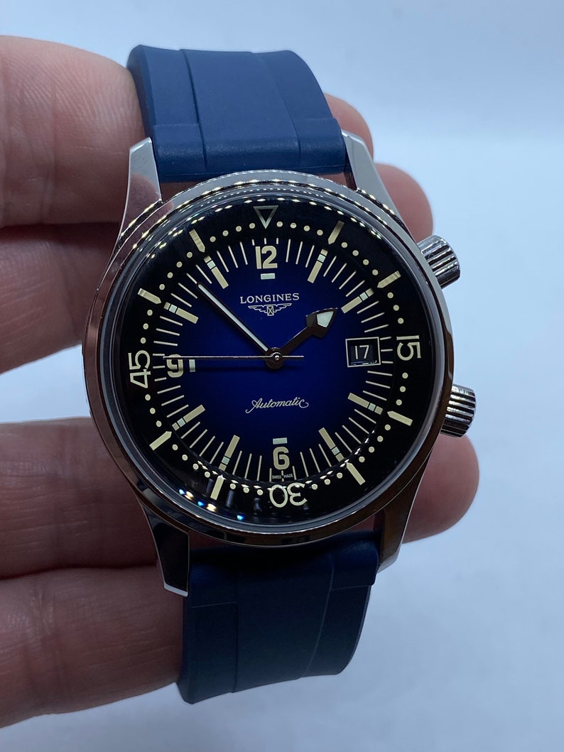May include: A silver Longines watch with a blue face and a black rubber strap. The watch has a date window at the 3 o'clock position and the words "Automatic" and "Longines" printed on the face.