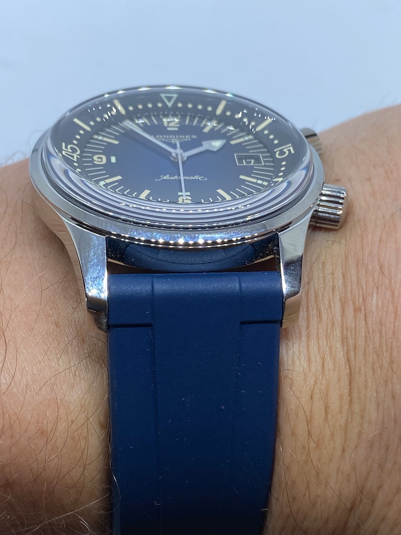May include: A silver Longines watch with a blue face and a blue rubber strap. The watch has a date window at the 3 o'clock position and a chronograph function.