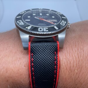 Puede incluir: Un reloj negro y plateado con una correa de caucho negro y costuras rojas. La esfera del reloj tiene un fondo negro con marcas blancas y una aguja de segundos roja. El reloj tiene las palabras "Seagull Master" en la esfera.