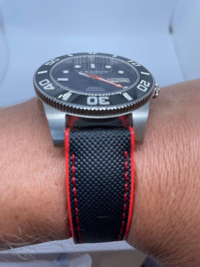 Puede incluir: Un reloj negro y plateado con una correa de caucho negro y costuras rojas. La esfera del reloj tiene un fondo negro con marcas blancas y una aguja de segundos roja. El reloj tiene las palabras "Seagull Master" en la esfera.