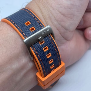 Peut inclure: Un bracelet de montre bleu marine avec des coutures orange et une boucle argentée. Le bracelet porte l'inscription "GOLBY" sur la boucle.