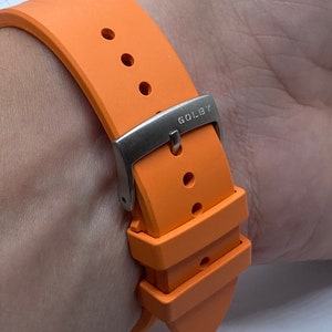 May include: Orange silicone watch strap with a silver buckle that says "GOLDBY".