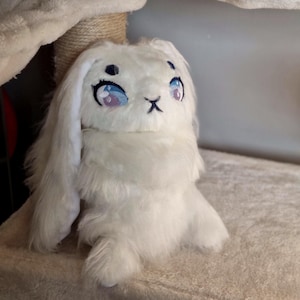 Può includere: Coniglio di peluche bianco con grandi occhi blu e naso rosa. Il coniglio ha orecchie lunghe e cadenti ed è seduto su una superficie marrone chiaro.