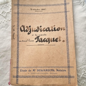 Può includere: Un documento d'epoca con il titolo "Adjudication" in scrittura ornata. La copertina è beige con bordi e testo decorativi blu, inclusa la data "14 Octobre 1906." Il documento è leggermente usurato, suggerendo l'età.