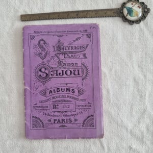 May include: A vintage purple booklet with ornate text and illustrations, including the words "SAJOU" and "PARIS." A metal ruler and a decorative metal bookmark with a bird design are also present. The booklet measures approximately 4 inches in length.