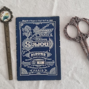 May include: An antique collection featuring a metal ruler with a bird design, a blue booklet with white text, and ornate scissors. The booklet reads "Ouvrages de Dames Maison Sajou Albums Dessins, Modeles, Fournitures".