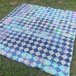Op de afbeelding: Een handgemaakte quilt met een ruitpatroon in tinten blauw, groen en bloemenprints. De quilt is uitgespreid op een grasachtig oppervlak en toont het ingewikkelde ontwerp en de kleurvariaties. Een traditioneel textiel.