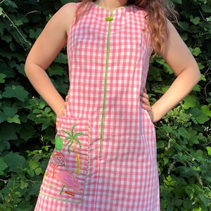 May include: Sleeveless pink and white gingham dress with a green zip and trim. The dress features a pocket with an embroidered flamingo, palm tree, and umbrella design. The dress is knee-length.