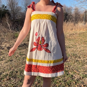 May include: A vintage-style, cream-colored dress with a large red floral applique. The dress features yellow and red patterned bands, and red bow accents on the straps. The model is wearing red flats. The dress is knee-length.