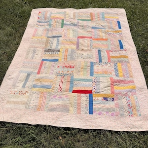 Puede incluir: Una colcha rectangular con un diseño de patchwork compuesto por varias tiras de tela de diversos colores y estampados, incluyendo flores, rayas y lunares. La colcha está bordeada por una tela de color rosa claro con un patrón sutil y repetitivo.