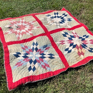 May include: A vintage quilt with four large star patterns in red, white, and blue on a light green background. The quilt has a red border and is displayed on green grass.