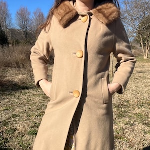 May include: A tan wool coat with a faux fur collar and large, round, wooden buttons. The coat has a classic design with a tailored fit, pockets, and a knee-length hem. The coat is worn outdoors on a sunny day.