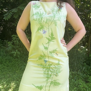 May include: A sleeveless, light yellow dress with a boat neckline. The dress features a green fern design with colorful butterflies. The dress is knee-length and appears to be made of a smooth fabric.